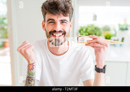 Junger Mann essen asiatische Sushi mit Stäbchen schreien stolz und feiern Sieg und Erfolg sehr aufgeregt, jubelnde Emotion Stockfoto