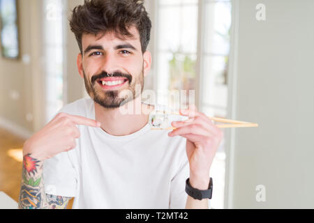 Junger Mann essen asiatische Sushi mit Stäbchen mit Überraschung Gesicht zeigenden Finger zu sich selbst Stockfoto