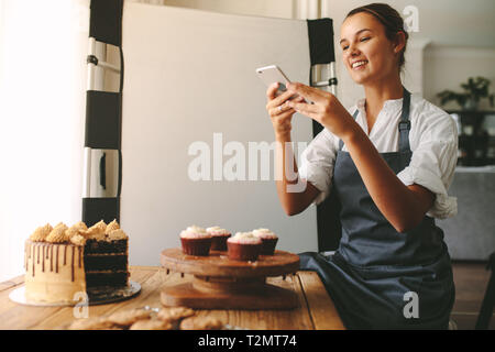 Glückliche Frau Koch ein Bild von Gebäck auf einem Holzbrett mit Smart Phone im Stehen in der Küche. Frau Baker, die Bilder von der De Stockfoto