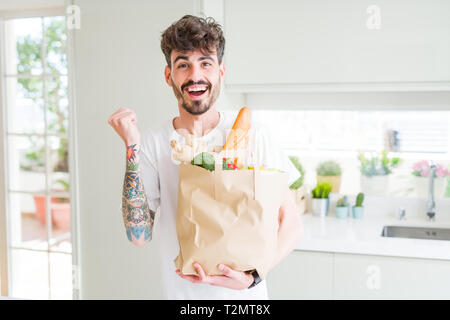 Junger Mann mit Papier Tüte frische Lebensmittel aus dem Supermarkt schreien stolz und feiern Sieg und Erfolg sehr aufgeregt, jubelnde Emotion Stockfoto