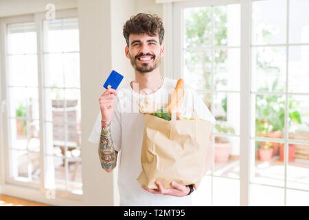 Jungen gutaussehenden Mann hält eine Papiertüte voller frische Lebensmittel zu Hause, die Kreditkarte als Zahlungsmittel metod Stockfoto