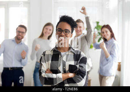 Fröhliche amerikanische junge Frau mit einer Gruppe von Multirassischen tausendjährigen fröhlichen Geschäft Leute im Hintergrund stehend zusammen lächelnd im Büro auf Stockfoto