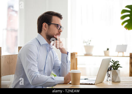 Nachdenklich tausendjährigen Kaufmann oder Unternehmer mit Brillen am Schreibtisch im Büro oder zu Hause arbeiten mittels Computer sitzen konzentriert. Th Stockfoto