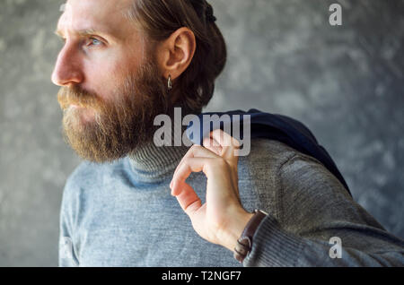 Mode Portrait von bärtigen Mann in Grau High Neck Pullover mit blauen Anzug Blazer hingen über seine Schulter auf grauem Hintergrund. Stockfoto