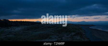 Panoramablick auf den Sonnenaufgang an der Cranbrook Warren, Anglesey. Sonnenaufgang über Snowdonia Berge in der Ferne, als Dawn bricht; dunkle Wolken, dunklen Küstenlinie. Stockfoto