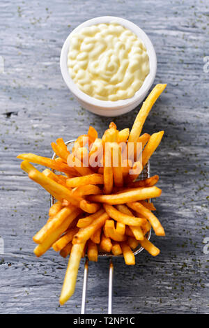 Hohe Betrachtungswinkel und einige leckere Pommes frites in einer metallischen Korb serviert, und ein weißer Keramik Schüssel Mayonnaise, auf einem grauen rustikalen Holztisch Stockfoto