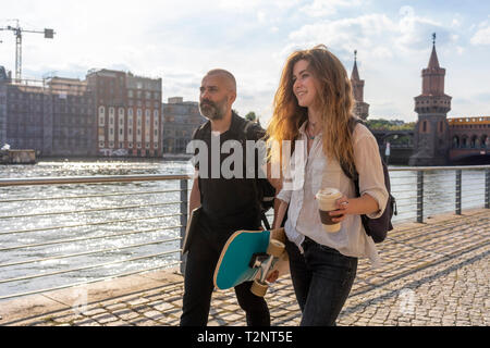 Mann und Frau Freund mit Skateboard auf Brücke, Fluss, Oberbaumbrücke und Gebäude im Hintergrund, Berlin, Deutschland Stockfoto