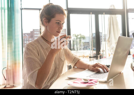 Junge Frau mit Laptop und Kaffee im Büro an einem sonnigen Tag Stockfoto