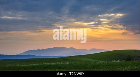 Sonnenuntergang in den Bergen. Grüne Felder und blaue Berge. Die Sonne versteckt sich hinter der orange Wolken. Kasachstan Stockfoto