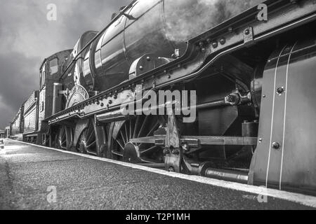 Schwarz-weiß, launisch, niedriger Winkel, Seitenhautnah von alten britischen Dampfeisenbahnrädern, die neben dem Bahnsteig abfahren. Dampflokomotiven in Großbritannien. Stockfoto