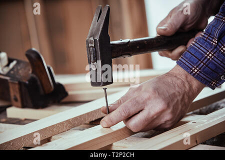 Tischler hämmern in einen Nagel mit Vintage Klauenhammer in Nahaufnahme auf seinen Händen und die Bretter, auf einer Werkbank Stockfoto