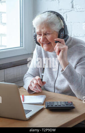 Eine alte Frau sitzt an einem Tisch vor einem Laptop mit Kopfhörern und schreibt in einem Notebook. Bildung für Erwachsene. Freelancer arbeiten. Pensionierte Frau ist Stockfoto