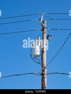 Ein elektrischer Transformator und Getriebe, Drähte, Kabel-TV und Telefon Kabel zu einem alten hölzernen Strommast mit einem tiefblauen Himmel Hintergrund geklebt. Stockfoto