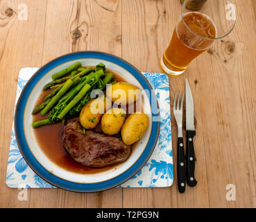 Eine gebratene Aberdeen Angus Rumpsteak Rindfleisch Steak serviert mit neuen Kartoffeln, Filets, Brokkoli und Spargel und Soße auf eine blau umrandete Platte auf einem Holztisch Stockfoto