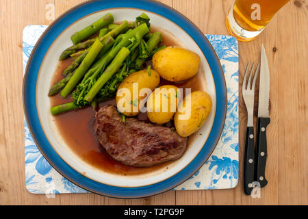 Eine gebratene Aberdeen Angus Rumpsteak Rindfleisch Steak serviert mit neuen Kartoffeln, Filets, Brokkoli und Spargel und Soße auf eine blau umrandete Platte auf einem Holztisch Stockfoto