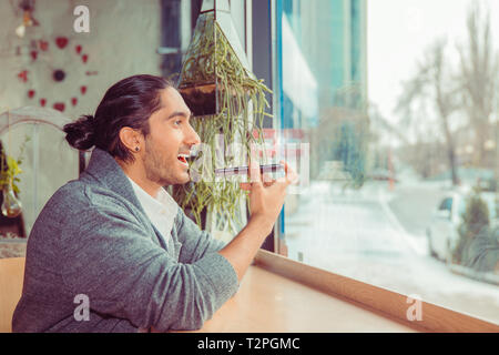 Mann mit Spracherkennung mit Smart Phone. Closeup Portrait hübscher Kerl tragen formalen weißes Hemd, grau Bluse sitzt in der Nähe der Fenster an einem Tisch in Li Stockfoto