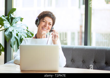 Die moderne Frau mittleren Alters tragen, halten Sie das Headset am Tisch sitzen vor dem Computer, Laptop an Sie Kamera suchen, glücklich lächelnd. Positive freel Stockfoto