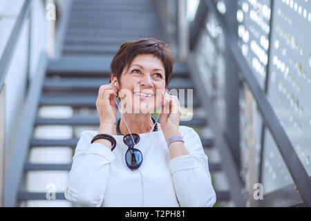 Stilvolle reife Frau in Kopfhörer Musik hören auf der Straße lächeln Treppen auf Hintergrund aufgeregt. Stockfoto