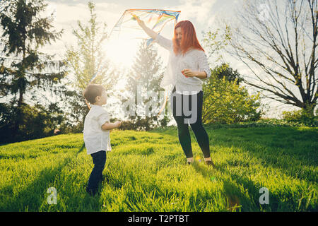 Schöne und stilvolle rothaarige Mama in eine weiße Bluse spielen mit ihren kleinen süßen Sohn in einem Sommer sonnigen Park mit einem Kite Stockfoto