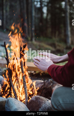 7/8 outdoor Bild des jungen Mannes Erwärmung seine Hände durch die in der Nähe von Feuer. Ansicht der Rückseite des Reisenden Mann im roten Mantel, in der Nähe von Lagerfeuer sitzen, nach Hiki Stockfoto