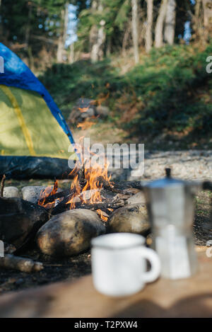 Unscharf Bild von Becher und Kaffeemaschine in der Nähe von Feuer in den Bergen. Selektiver Fokus auf ein Lagerfeuer mit unscharfem Hintergrund. Reisen, Lifestyle Konzept Stockfoto