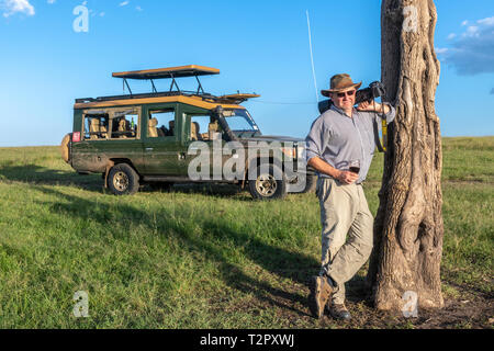 Ein Fotograf stellte hält seine Kamera und ein Drink vor einem Spiel Viewer in Masai Mara National Reserve, Kenia Stockfoto