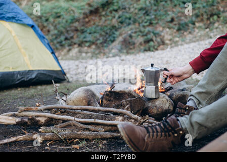 7/8 outdoor Bild des jungen Mannes explorer warme Getränke in den Bergen. Reisende Mann sitzt in der Nähe auf dem Hintergrund camping Zelt, Kochen am Lagerfeuer Stockfoto