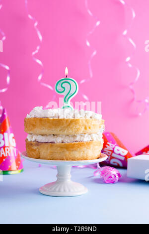 Eine festliche Biskuitteig mit weißer Creme für einen Geburtstag, auf einem Rosa und blauen Hintergrund. Lametta, waverki und Boxen von Geschenken. Fragezeichen Kerze am Stockfoto