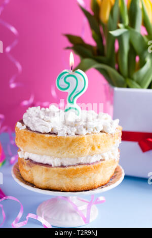 Eine festliche Biskuitteig mit weißer Creme für einen Geburtstag, auf einem Rosa und blauen Hintergrund. Lametta, waverki und Boxen von Geschenken. Fragezeichen Kerze am Stockfoto