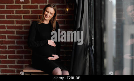 Junge hübsche schwangere Frau in schwarzem Kleid im Studio-loft Innenraum Stockfoto