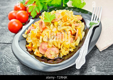 Kohleintopf mit Würstchen in einer schwarzen Platte, Handtuch, Tomaten, Petersilie und Gabel auf dem Hintergrund der Holzbretter Stockfoto