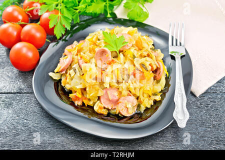 Kohleintopf mit Würstchen in einer schwarzen Platte, Serviette, Tomaten, Petersilie und Gabel auf einem dunklen Holzplatte Hintergrund Stockfoto