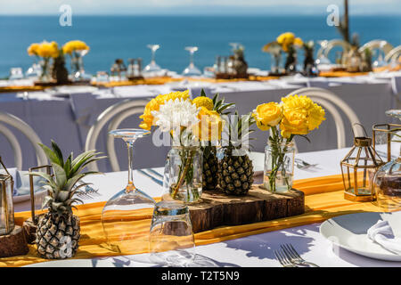 Weiß gedeckten Tischen für Hochzeit Abendessen mit gelben Rosen, Chrysanthemen, Ananas und Glas Lampen eingerichtet. Blick auf den Ozean Stockfoto
