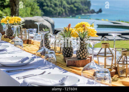 Weiß gedeckten Tischen für Hochzeit Abendessen mit gelben Rosen, Chrysanthemen, Ananas und Glas Lampen eingerichtet. Blick auf den Ozean Stockfoto