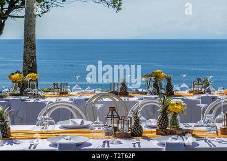 Weiß gedeckten Tischen für Hochzeit Abendessen mit gelben Rosen, Chrysanthemen, Ananas und Glas Lampen eingerichtet. Blick auf den Ozean Stockfoto