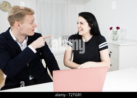 Vorgesetzten arbeitet mit unterordnen. Blonder mann Punkte Finger auf schwarzhaarige Frau, sie lacht Stockfoto