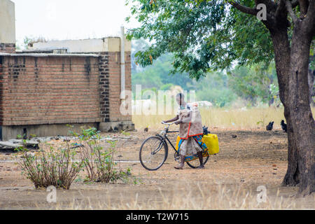 Malawische Frau drücken Fahrrad mit leeren Wasserbehälter an der Pumpe geladen, bevor ein verfallenes Gebäude Stockfoto