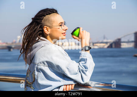 Eine schöne junge Kaukasische Mädchen mit Dreadlocks und rasiert Tempel ist das Trinken Kaffee aus einer grünen - Zeit Schale, während sie durch den Fluß. Mindfuln Stockfoto