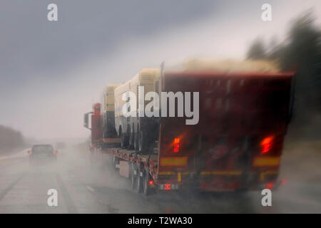 Das Fahren auf der Autobahn bei Regen und vorbei an einem Semi Truck mit einer Last von Maschinen, Regenwasser, Spritzwasser, verschwommenes Sehen. Stockfoto