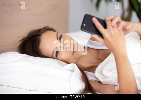 Junge attraktive Frau liegend auf Bett holding Telefon Nachricht lesen Stockfoto