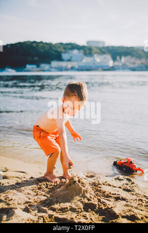 Thema Bau- und Schwerindustrie. Abstraktion Kind Junge beim Spielen im Sand in der Nähe des Flusses im Sommer Spielzeug rot Traktormodell, Bagger machi Stockfoto