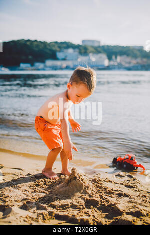 Thema Bau- und Schwerindustrie. Abstraktion Kind Junge beim Spielen im Sand in der Nähe des Flusses im Sommer Spielzeug rot Traktormodell, Bagger machi Stockfoto