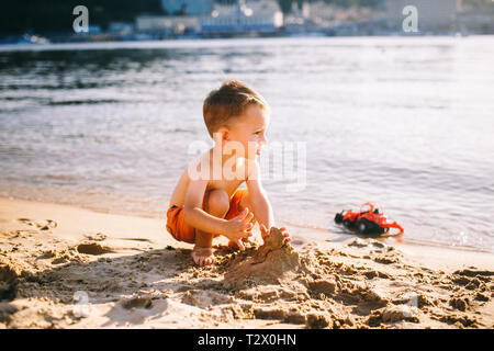 Thema Bau- und Schwerindustrie. Abstraktion Kind Junge beim Spielen im Sand in der Nähe des Flusses im Sommer Spielzeug rot Traktormodell, Bagger machi Stockfoto