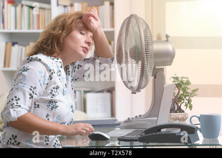 Frau leidet unter der Hitze, während der Arbeit im Büro und versucht durch den Lüfter zu kühlen Stockfoto