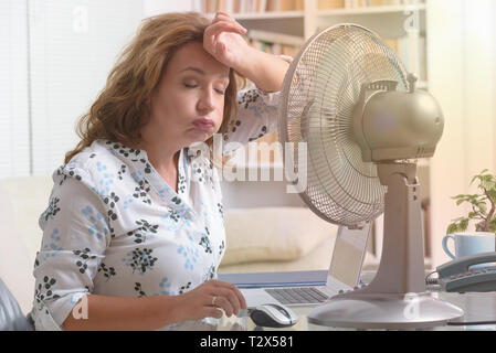 Frau leidet unter der Hitze, während der Arbeit im Büro und versucht durch den Lüfter zu kühlen Stockfoto