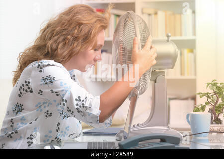 Frau leidet unter der Hitze, während der Arbeit im Büro und versucht durch den Lüfter zu kühlen Stockfoto