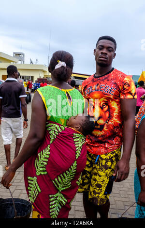 SEKONDI TAKORADI, GHANA - 10. APRIL 2018: Junges Paar in bunte Kleidung mit schlafenden Baby an belebten Bosomtwi Sam Fischer Hafen Fischmarkt. Lion o Stockfoto