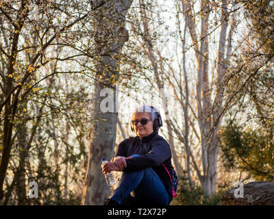 Eine ältere Frau mit weißen Haaren Musikhören mit Kopfhörern und einem Kunststoff Flasche Wasser in der Hand. Stockfoto