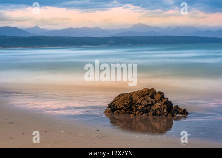 Verträumt Wellen in Zeitlupe in Plettenberg Bay Strand bei Sonnenuntergang brechen, mit Hügeln in der Ferne und ein Stein im Vordergrund spiegelt sich in der ... Stockfoto