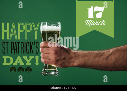 7/8-Ansicht der Mann hält ein Glas Bier mit Schaum in der Nähe von Happy st patricks day Schriftzug auf grünem Hintergrund Stockfoto